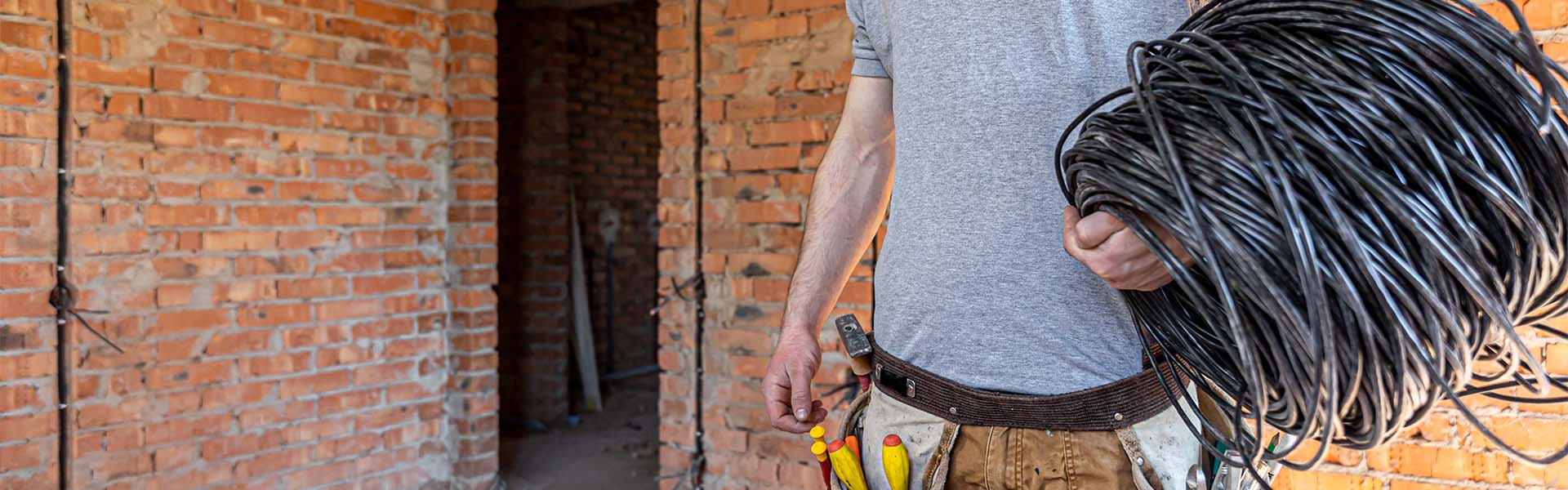 An electrician in a hard hat looks at the wall while holding an electric cable.